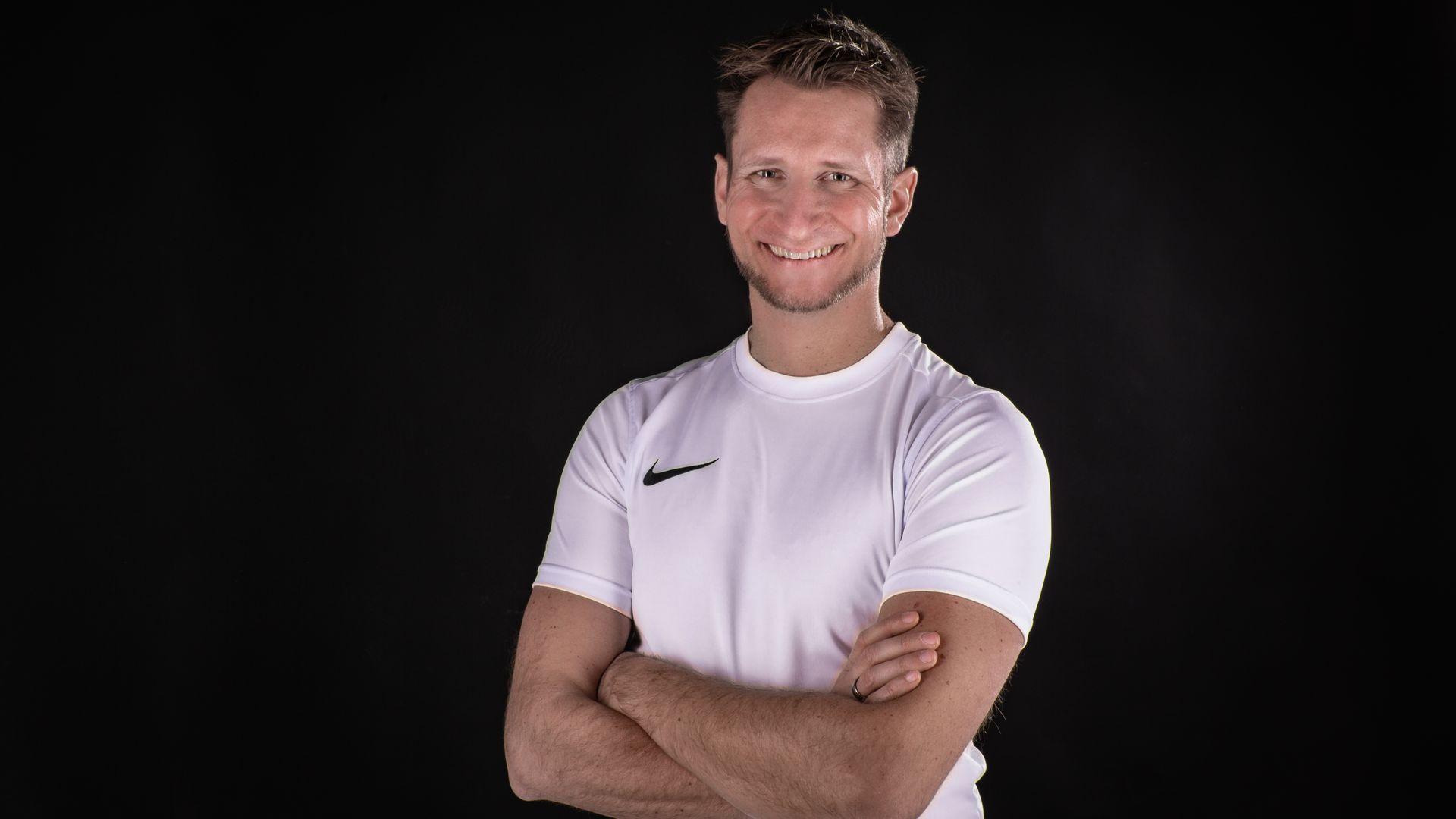 Man in a white shirt with crossed arms, smiling against a dark background.